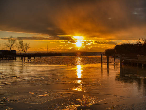 A tranquil winter sunset casts a golden glow across a frozen lake, its surface fragmented into shimmering ice patterns. The silhouette of trees and a docked sailboat add charm to the serene scene. Dark clouds frame the horizon, accentuating the vivid oranges and blues of the sky. A lakeside structure and distant mountains provide a peaceful backdrop, embodying the quiet beauty of nature in winter.