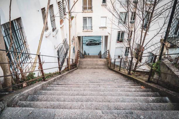 A concrete staircase winds down between two buildings, revealing a tranquil street below. The surroundings are characterized by white facades and slender trees, hinting at a residential neighborhood. The steps are bordered by metal railings, and the street is mostly devoid of traffic, evoking a sense of calm and solitude. Signs of urban living blend with nature, creating a peaceful atmosphere for those walking down the path.