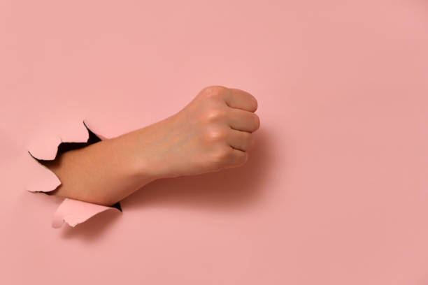 istock Human hand showing clenched fist through torn pink paper background. Studio conceptual photography. Design for poster, banner, wallpaper, and header. 2193546247