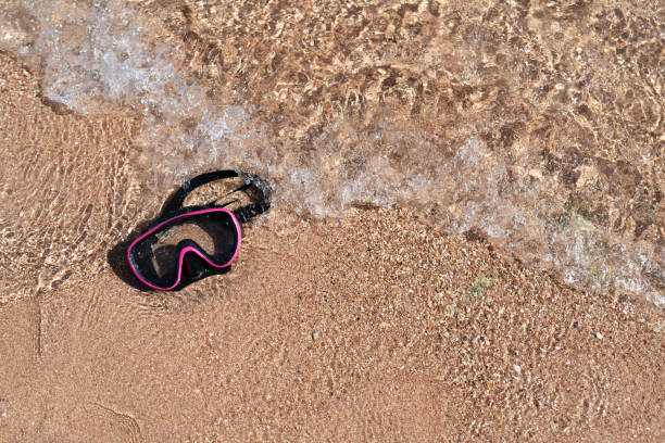 A diving mask lies on a sandy beach and is washed by waves of water. Free space for your materials. A photo with a high resolution.