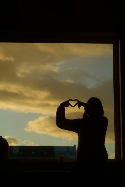 Silhouette of a young woman making a heart shape with her hands in front of a window at sunset