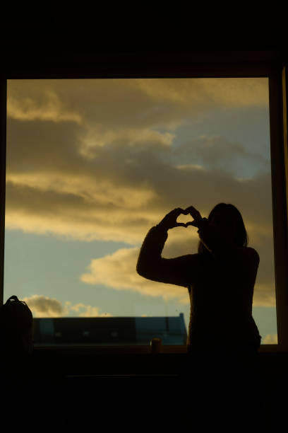 Female silhouette making heart shape with hands by the window at sunset