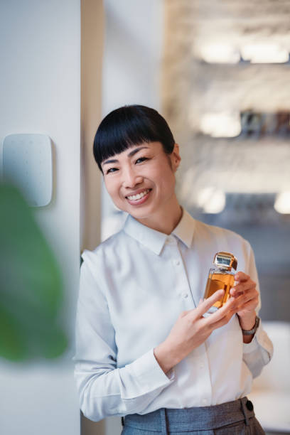A cheerful woman holding a bottle of perfume while leaning against a wall in a bright space. She appears relaxed and content, showcasing the perfume with an inviting and pleasant demeanor.