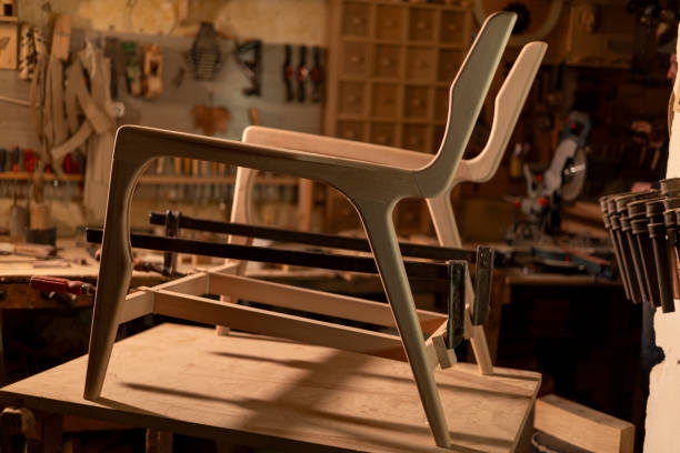 Modern design wooden chair frame resting on workbench in carpentry workshop, surrounded by tools and wood pieces, illuminated by warm light