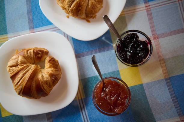 Two golden croissants served with jars of delicious dark berry and orange jams on a colorful checkered surface. A cozy breakfast setting suggesting warm mornings and delightful treats.