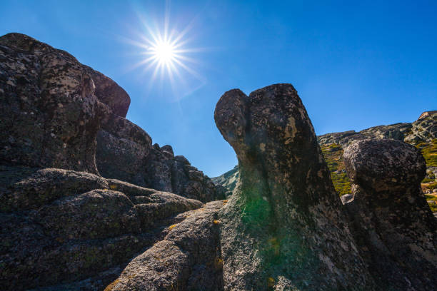 Jagged rock formations rise against a bright blue sky with a shining sun, creating a dramatic and powerful natural scene