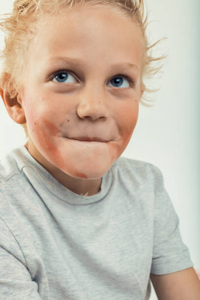 Close-up portrait featuring a blond boy with bright blue eyes, gazing upward with chocolate smeared around his mouth, set against a clean white background