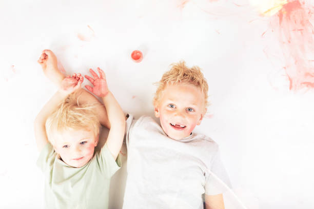 Two blonde brothers lying on a white background, joyfully covered in pink paint, smiling at the camera and radiating happiness and playful creativity in their messy fun