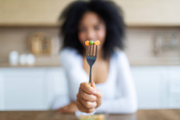 Young woman eating vegetables, showing fork with green peas and carrot