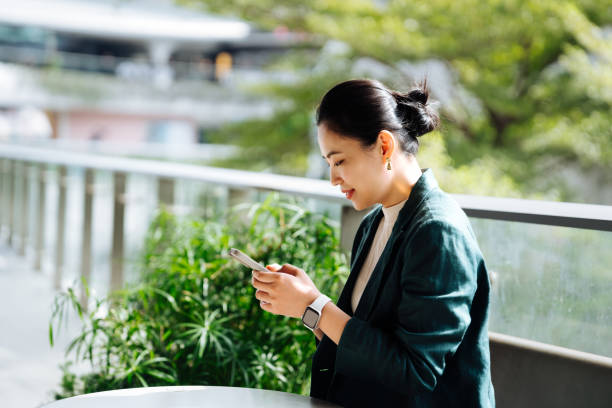 istock Asian business woman using smartphone working on digital tablet in business park. Taking a break from work. Remote working with flexibility. Working outdoors with technology. Business lifestyle. Work-life balance and wellbeing concept . 2193641523