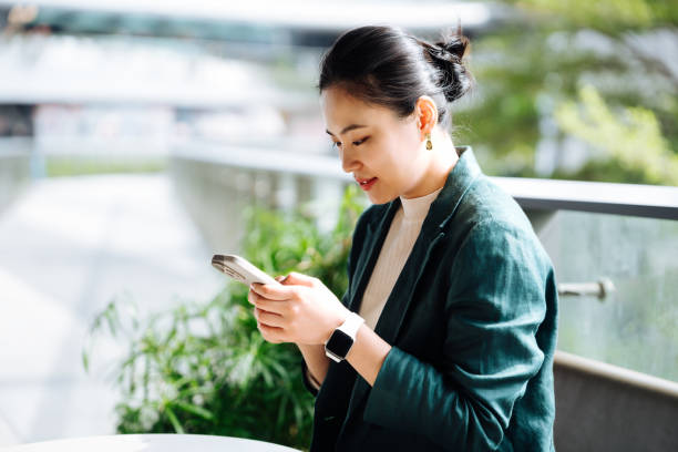 Asian business woman using smartphone working on digital tablet in business park. Taking a break from work. Remote working with flexibility. Working outdoors with technology. Business lifestyle. Work-life balance and wellbeing concept .