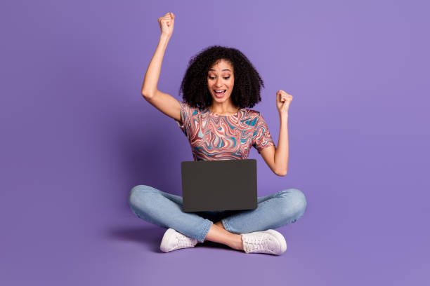 istock Young woman celebrating success while using laptop against vibrant violet background, expressing excitement and happiness 2193662240