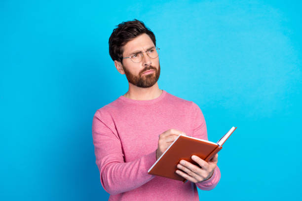 istock Thoughtful bearded man in glasses writing notes in a journal, dressed in pink against a vibrant blue background, conveying a concept of creativity and contemplation 2193667353