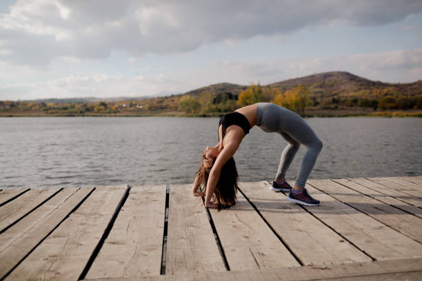 A woman performing a yoga backbend exercise on a wooden dock, surrounded by tranquil natural scenery with a calm lake and autumn foliage in the background, promoting flexibility, fitness, and connection to nature.