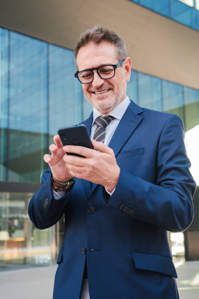 istock Vertical. Corporate business man smiling using a smartphone at workplace. Real successful executive male or ceo watching a finance app on his cellphone. Businessperson holding a mobile phone smiling 2193678302