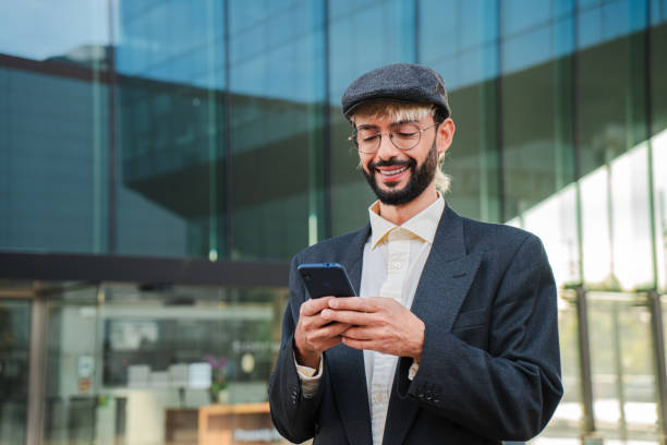 istock Young adult man using a cellphone app at workplace. Bearded cool guy having fun with a smartphone device. Caucasian positive male texting and sending messages with a mobile phone. Cool businessman 2193682188
