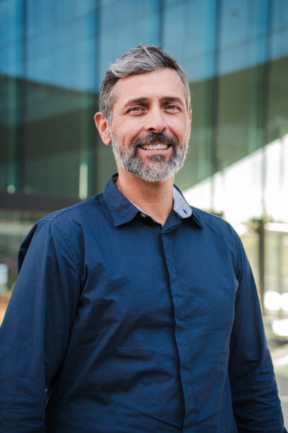istock Vertical Bearded mature man smiling and looking at camera. Middle aged male entrepreneur staring front with friendly and confident expression. Individual portrait of a mid adult guy wearing blue shirt 2193685438