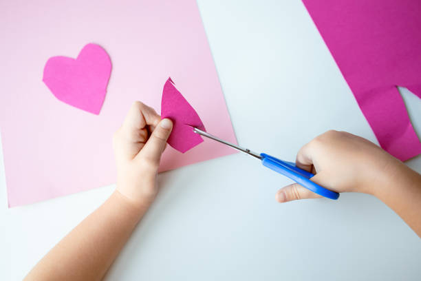 istock Child cutting pink paper heart with scissors on white table 2193689588