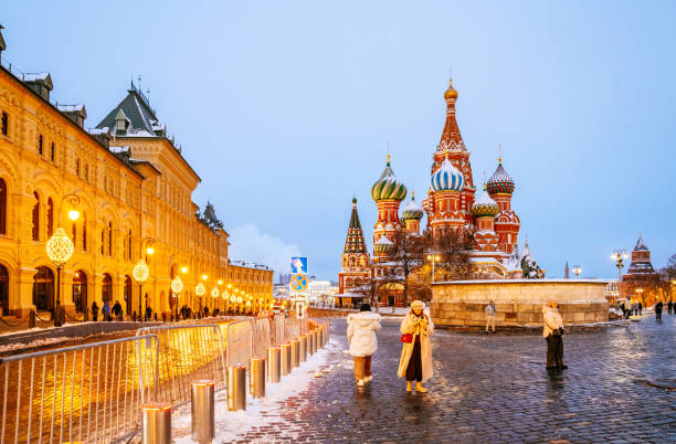 St. Basil’s Cathedral stands prominently with its colorful domes and intricate architecture. Snow-dusted cobblestones reflect soft festive lights, while visitors explore Red Square during a winter evening.