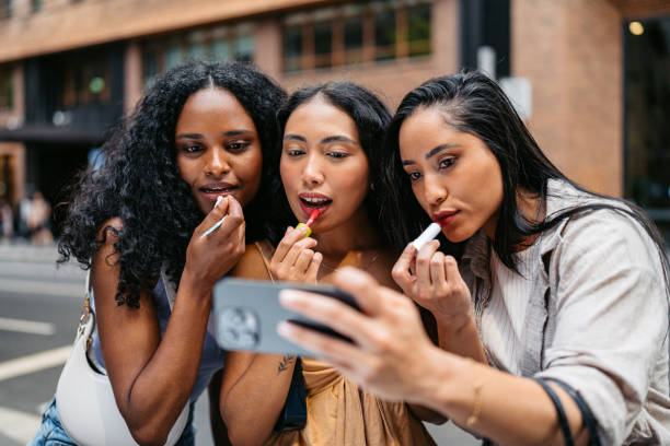Three Young Female Friends Applying Lipstick While Looking At Their Reflection On The Phone Screen On The Street In Sydney In Australia Three young female friends applying lipstick while looking at their reflection on the phone screen on the street in Sydney in Australia. three way kiss stock pictures, royalty-free photos & images