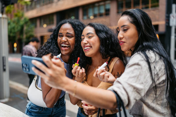 Three Young Female Friends Applying Lipstick While Looking At Their Reflection On The Phone Screen On The Street In Sydney In Australia Three young female friends applying lipstick while looking at their reflection on the phone screen on the street in Sydney in Australia. three way kiss stock pictures, royalty-free photos & images