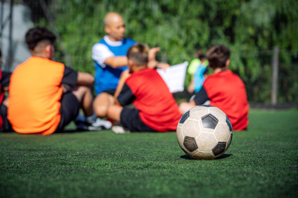 Young children grouped on a soccer field, listening attentively to their coach, fostering teamwork and learning sporting strategies. A soccer ball rests in the foreground on the lush green field.