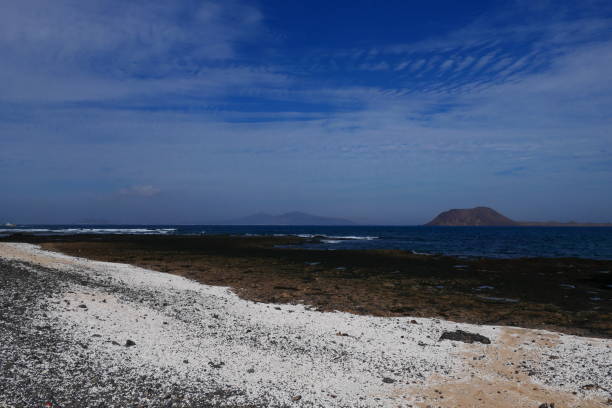 Calm, turquoise waters gently lap the sandy shore of Lanzarote, with Lobos Island visible in the distance, offering a serene and picturesque view