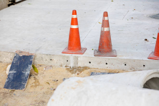 Yellow caution tape and orange traffic cones cordon off a damaged pillar and rubble on a street, indicating a construction or accident zone