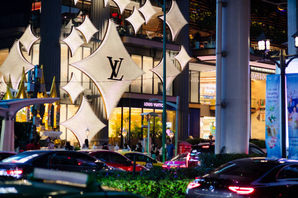 Illuminated facade of Gaysorn Tower (Gaysorn Village) in Bangkok Lumpini located at Phloen Chit Road. View over street with some traffic. Outside os golden shiny futuristic decor for Louis Vuitton. A Louis Vuitton store is behind in building. People and tourists are walking along sidewalk. In right area is entrance to mall complex. Street lanes are separated by a hedge.
