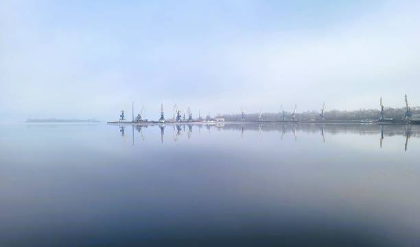 The photograph depicts a row of port cranes standing along the shore and reflected in the calm, foggy water. The fog creates soft, diffused lighting and gives the landscape a mysterious atmosphere. In the background, the outlines of the coastline are barely visible