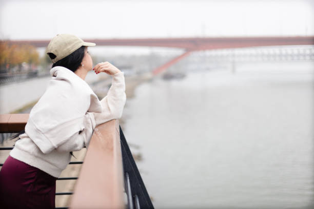 A thoughtful woman dressed in casual attire leans on a riverside railing overlooking a bridge. The surrounding ambiance is calm, showcasing a serene waterfront during the transitional season of autumn.
