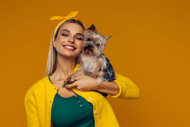 A beautiful young woman with bright, expressive eyes and a radiant smile holds her Yorkshire Terrier close. She wears a yellow headband and a matching outfit, complementing the warm, orange background. Her cheerful expression and the dog's calm demeanor create a heartwarming and joyful atmosphere, perfect for themes related to pets, companionship, and happiness.