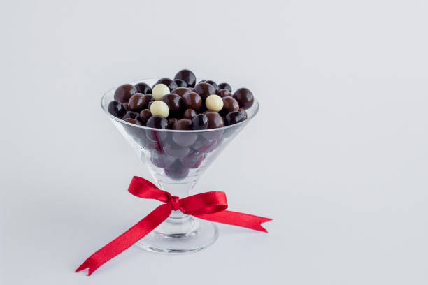 Elegant dark ball chocolates in glass candy bowl at white surface with red bow and copy space.