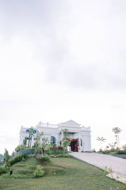 istock White colonial-style building with a lush garden and pathway under a bright, cloudy sky. 2193773388