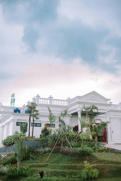 istock White colonial-style building with a lush garden and pathway under a bright, cloudy sky. 2193773479