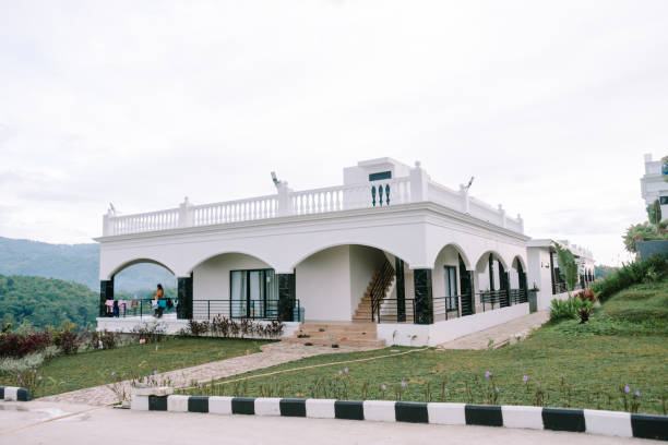 A luxurious white colonial-style building surrounded by vibrant greenery and a landscaped lawn, with a clear pathway leading to its entrance under a bright, cloudy sky