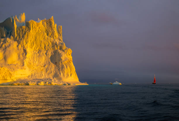 red sailboat sailing among floating yellow icebergs hit by setting sun in Disko Bay during polar summer midnight solar season. Ilulissat, Greenland