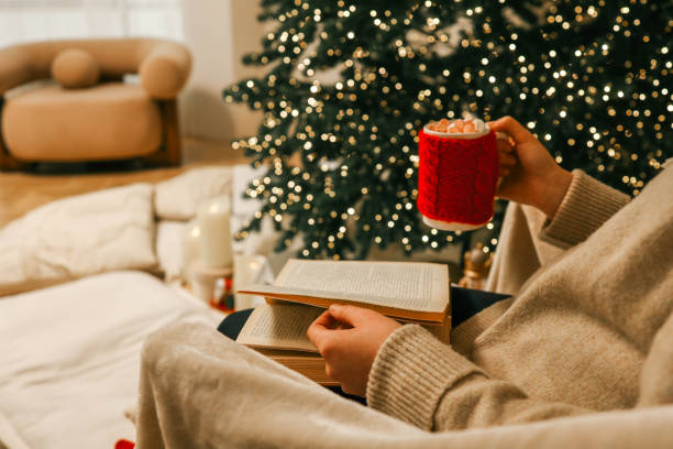 istock A cozy holiday scene featuring a person reading an open book while holding a festive red mug filled with marshmallows 2193814009