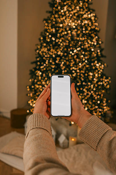 istock Person holding a smartphone with a blank screen in a cozy holiday setting. A decorated Christmas tree sparkles warmly in the background, creating a festive and serene atmosphere 2193814202
