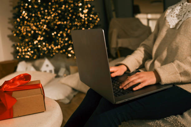 istock Hands typing on a laptop in a cozy festive setting. A beautifully decorated Christmas tree glows in the background, while a wrapped gift box with a red ribbon sits on a nearby table 2193814212