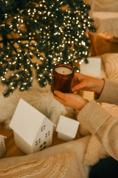 istock Hands holding a jar candle in a cozy Christmas setting with glowing candles, a decorated tree, and soft textures. Warm and festive atmosphere ideal for holiday relaxation and celebration 2193814230