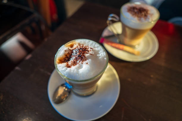 Two warm coffee drinks are placed on small white plates on a dark wooden table, creating a cozy atmosphere. The drinks are topped with a generous layer of frothy milk and a sprinkle of cocoa powder, inviting indulgence. Dim lighting enhances the intimate setting of the cafe, allowing customers to enjoy their beverages while possibly engaging in conversation or relaxing. The ambiance suggests a perfect spot for coffee lovers.