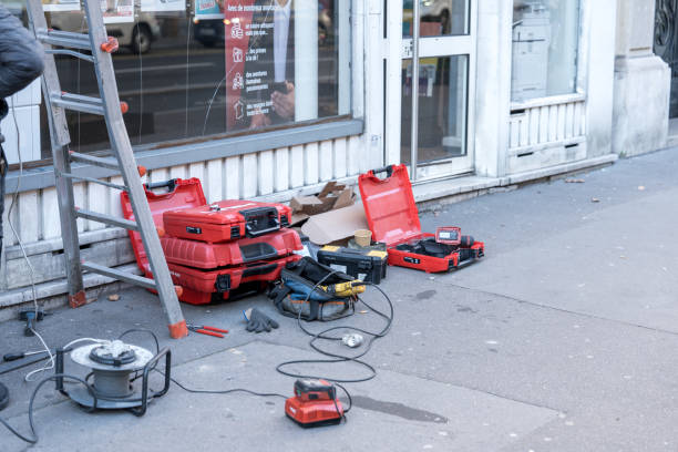 On a busy street in Paris, a work site is bustling with activity. Various construction tools and equipment are neatly arranged on the sidewalk. Bright red tool cases and power devices lay scattered, indicating ongoing renovations or repairs. A ladder leans against a building alongside the tools, signifying a hands-on project. The winter atmosphere is palpable as pedestrians navigate around the work area, showcasing urban life in January 2025.