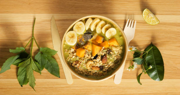 Verdant green smoothie bowl brimming with spinach, crowned with banana, mango, granola, chia seeds, nuts on rustic wooden surface with botanical garnish