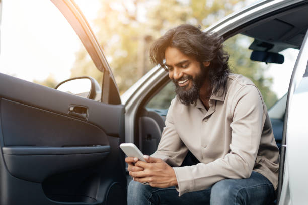 istock Young man sitting in car doorway, smiling while using smartphone outdoors during sunny day 2193850916