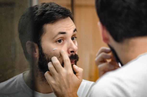 istock A man is applying moisturizer to his face while looking at himself in the mirror - stock photo 2193852036