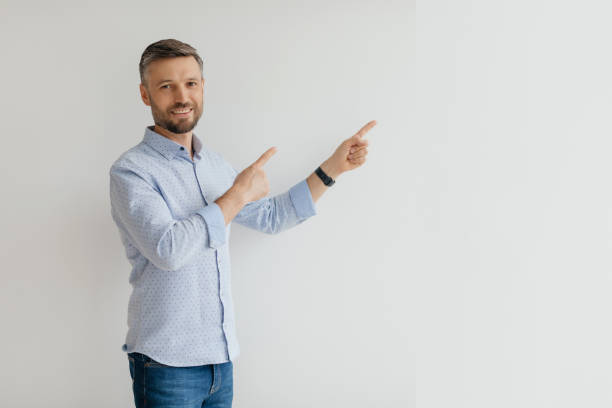 istock Man in a light blue shirt smiling and pointing at an empty wall in a bright, minimalistic interior during the daytime 2193867698