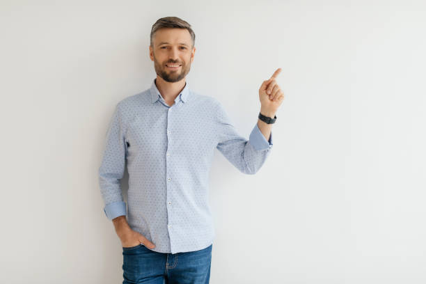 istock Smiling man in a blue shirt points upward against a plain white wall in a bright, well-lit indoor setting 2193868797