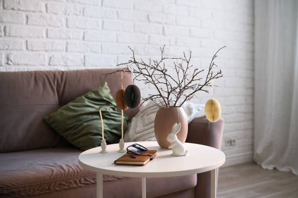 A beautifully cozy living room with modern easter home decor that includes vase with dry branches and eggs, notebook, glasses, ceramic rabbit, candles on coffee table.