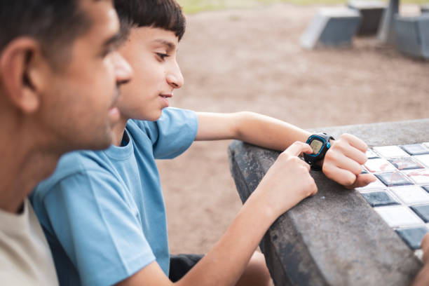 A father and son spend time bonding at a park, using smartwatch, sharing a relaxed moment, and nurturing their relationship as they enjoy the outdoor environment on a sunny day.